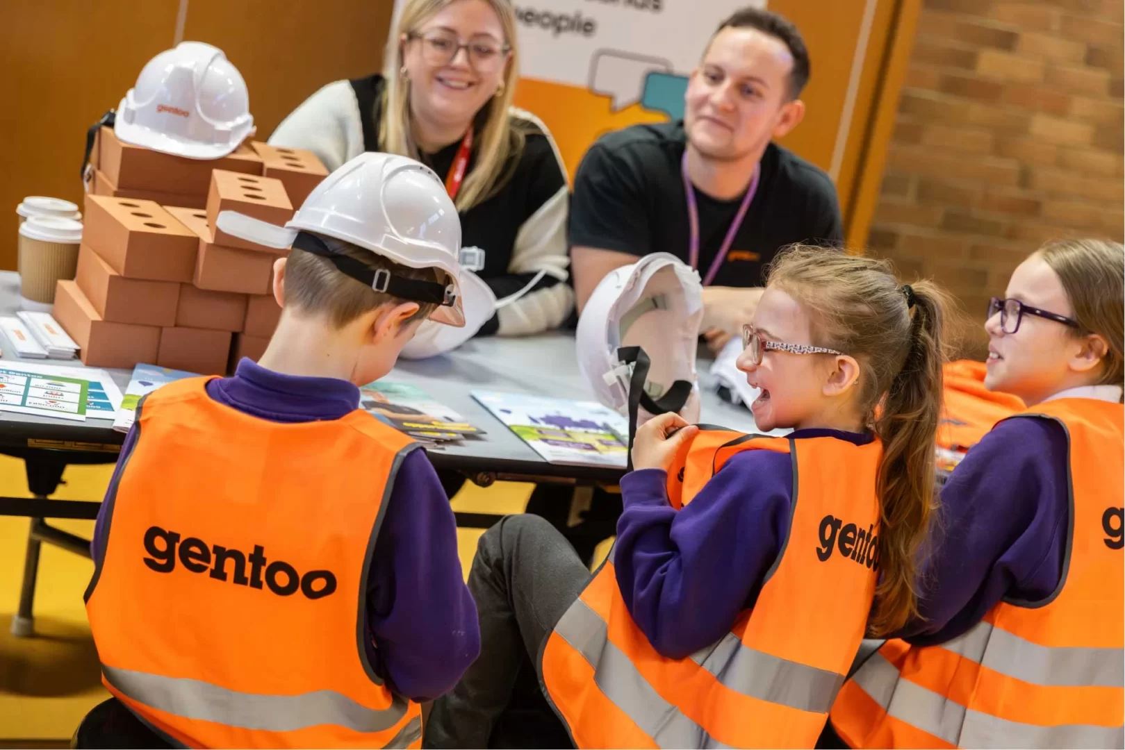 Group of young pupils at the forefront sat at a table, smiling whiling trying on safety gear such as hard hats and high-vis' jackets, in the background are staff from Gentoo smiling