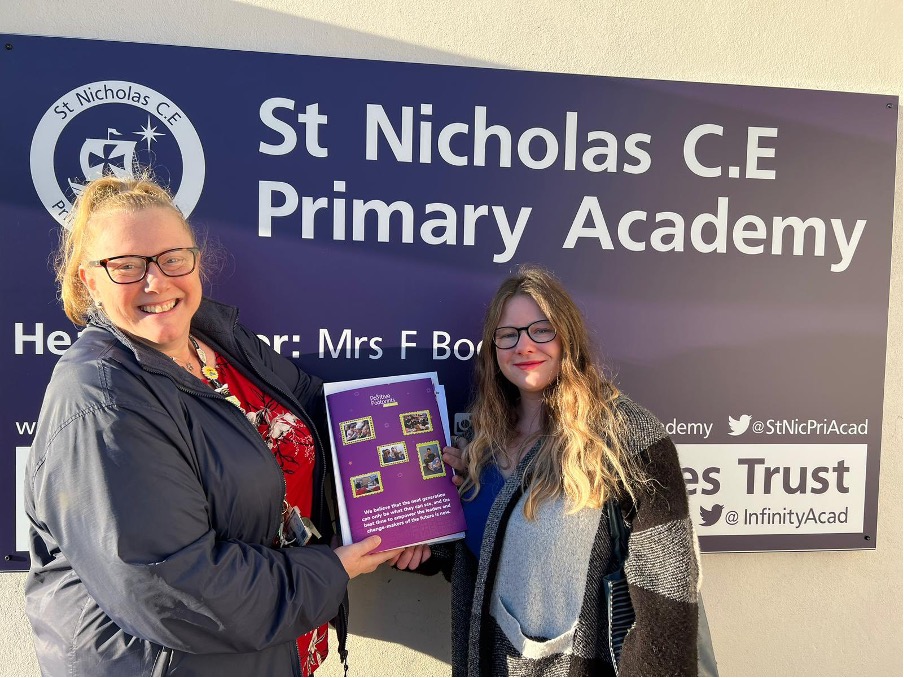 Positive Footprint Staff meeting with Teacher, smiling infront of School board that reads "St. Ncholas C.E Primary Academy"