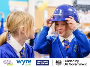 Two 10 year old girls in blue school uniform smiling at each other, one on is trying on a hard hat. Logo's pictured at the bottom represent Positive Footprints, Wyre Council, Leveling up and Funded by UK Government