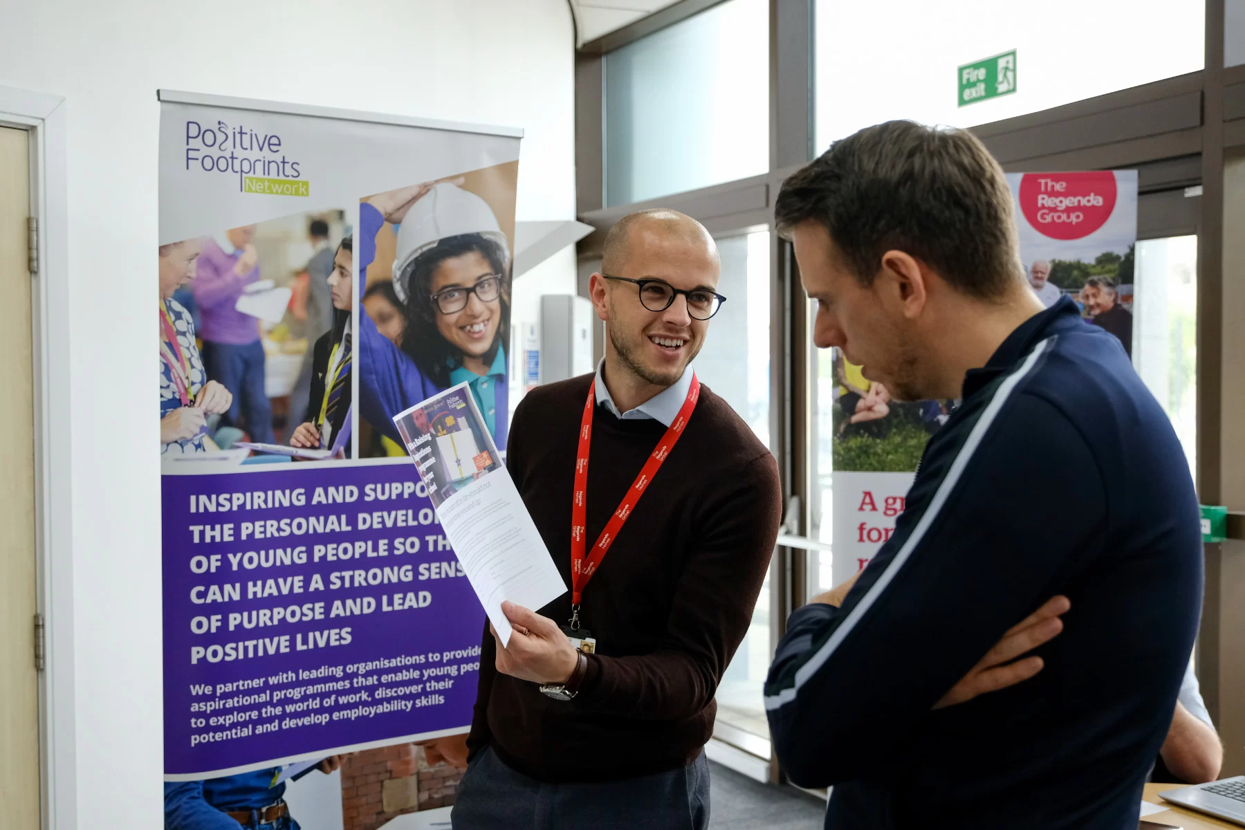 Josh Cliff, head of service at Positive footprints shows a brochure to a teacher in front of a pull up banner featuring young people learning.